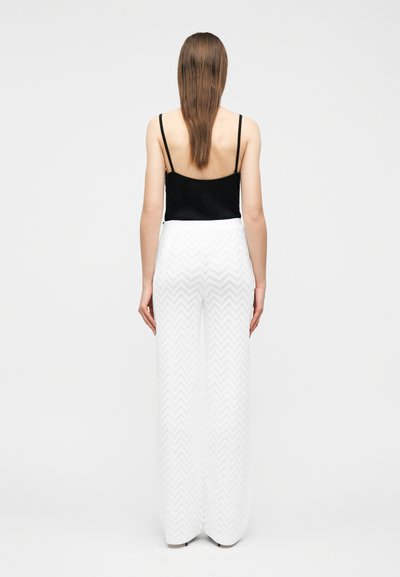 Γυναίκα στέκεται γυρισμένη με την πλάτη φορώντας μαύρο τοπ με λεπτούς ιμάντες και ψηλόμεσο άσπρο παντελόνι με σχέδιο ζιγκ ζαγκ.