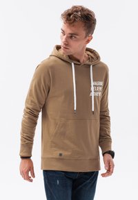 Felpa con cappuccio marrone e lacci bianchi, tasca frontale e testo stampato: "IMMAGINA CREDI RAGGIUNGI." Indossata con jeans blu strappati.