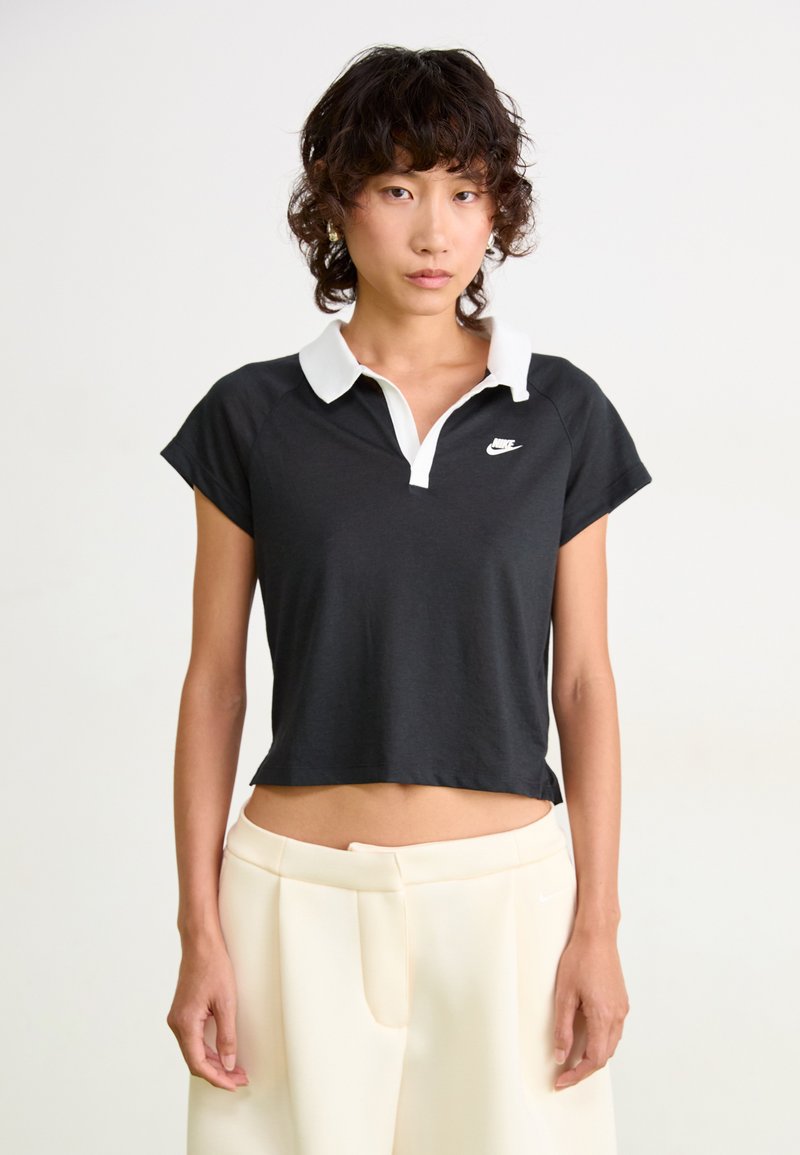 Čierna skrátená polo košeľa s bielym golierom a logom Nike, s krátkymi rukávmi a voľným strihom, nosená s krémovými nohavicami.