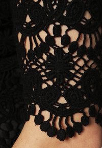 Tissu en dentelle noire avec des motifs floraux complexes et des designs tourbillonnants. Présente des accents décoratifs de pompons le long du bord.
