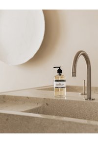 Bouteille à pompe en verre transparent contenant du savon liquide, étiquetée "Polivre Noir Frais", placée sur un comptoir beige texturé à côté d'un robinet en métal brossé.