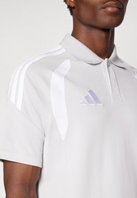 Ανοιχτό γκρι πόλο με υφή υφάσματος, λευκές λεπτομέρειες στους ώμους και διακριτικό μωβ λογότυπο Adidas στο στήθος. Κλασικός σχεδιασμός γιακά.