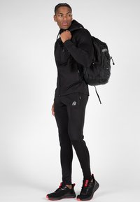 Juodas sportinis džemperis su kapišonu ir kelnes; elegantiškas dizainas su šoninėmis kišenėmis; derinamas su juodais ir raudonais sportbačiais; audinys atrodo lygus ir prigludęs.