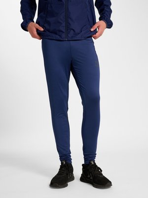 Person iført marineblå træningsbukser, sorte løbesko og en matchende marineblå jakke, stående mod en ensfarvet baggrund.