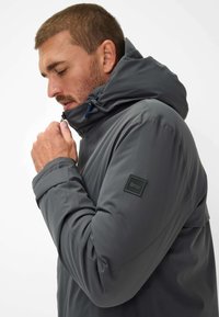 Uomo in giacca grigia con colletto alzato, mentre la chiude, visto di lato con focus sulla parte superiore del corpo e sulla toppa del braccio.