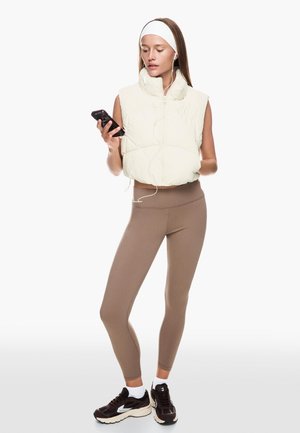 Witte, puffy mouwloze vest over bruine leggings; draagt zwarte sportschoenen en een witte haarband; houdt een telefoon vast met oordopjes.