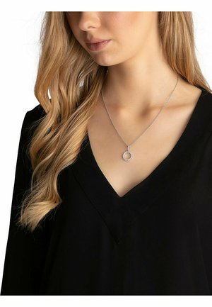 Sølvhalskæde med en cirkulær vedhæng pyntet med små klare sten, båret med en sort V-hals bluse. Glat metal kæde.