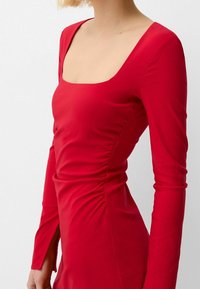 Rød langærmet kjole med firkantet halsudskæring, der har sammensyninger på siden og en tætsiddende silhuet. Glat, strækbart stof.