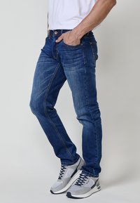 Jeans de mezclilla azul con un corte ajustado, ligera decoloración y costuras en contraste. Llevados con zapatillas atléticas grises con acentos de malla y ante.