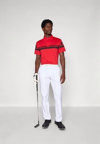 Ung man står med en golfklubba, iklädd en röd polo med svarta ränder, vita byxor, svarta skor och en vit golfhandske.