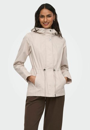 Femme portant une veste à capuche beige avec poches avant et cordons de serrage, associée à un pantalon marron foncé, souriante avec les mains dans les poches.