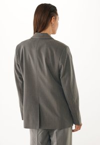 Blazer gris ajustado con una textura suave, abertura trasera sencilla y solapas de muesca. Presenta sutiles rayas finas. Vista trasera lisa.