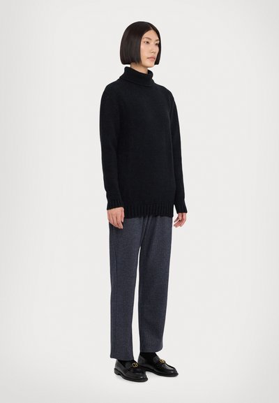 Svart oversized turtleneck-genser i myk strikk, sammen med grå skreddersydde bukser. Svart loafers med gullakenter sitter på føttene.