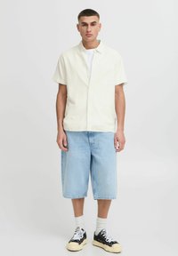 Chemise à manches courtes, jaune clair, avec un motif texturé, associée à un short en denim léger bleu clair et des baskets noires.