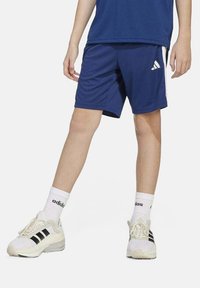 Pantalones cortos deportivos azules con rayas blancas a los lados, combinados con zapatillas blancas que tienen rayas negras y calcetines blancos con canalé.