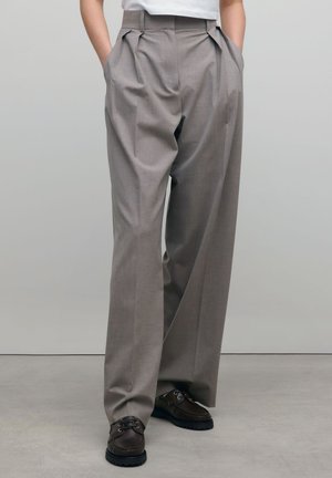 Personne portant un pantalon ample gris plissé avec les mains dans les poches et des chaussures à lacets marron foncé, debout sur un sol gris.