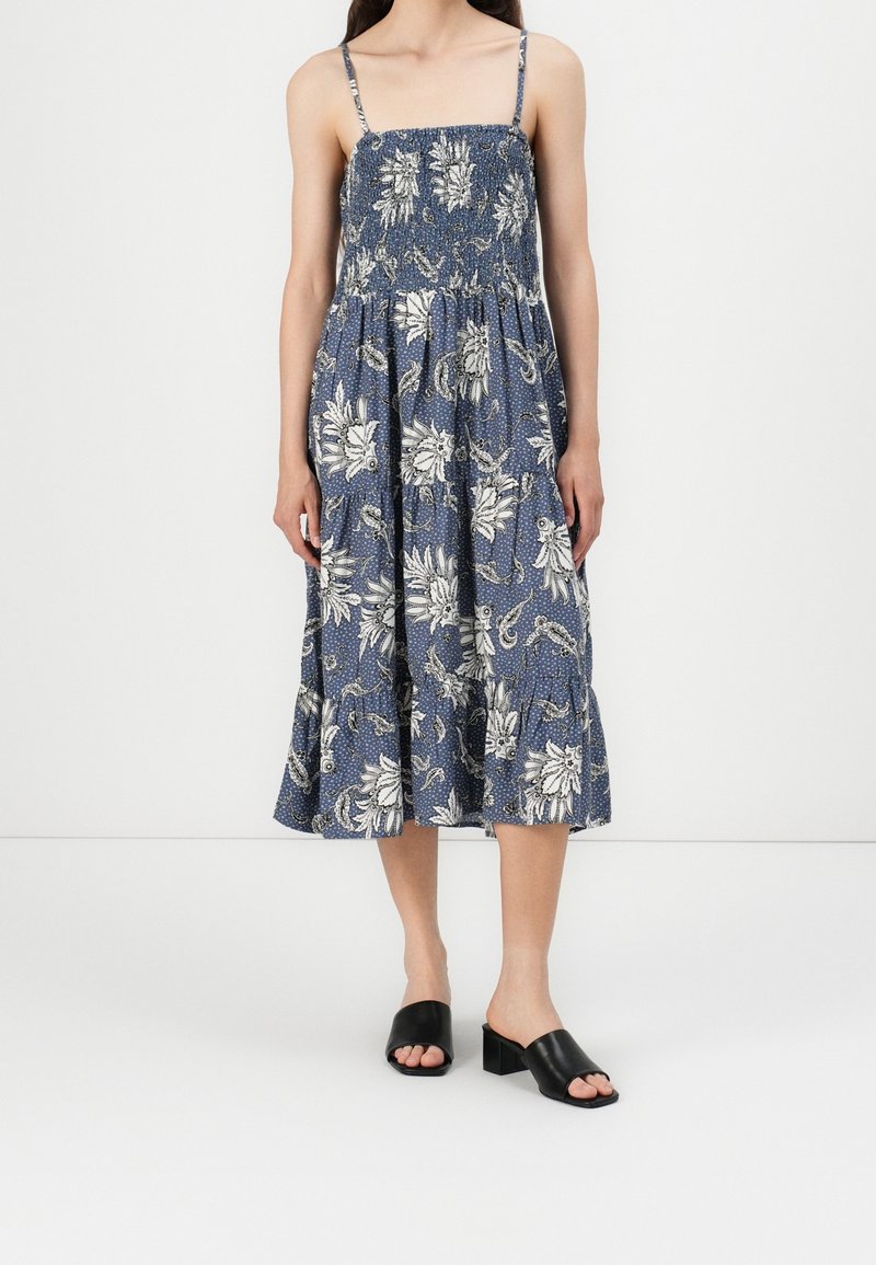Femme portant une robe midi bleue avec imprimé floral blanc et fines bretelles, associée à des sandales noires à talons blocs ouvertes à l'avant.