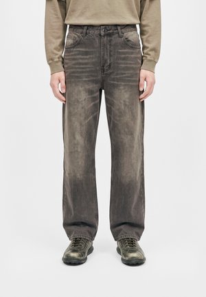 Un mannequin porte un jean ample et délavé gris anthracite avec des poches avant, un sweat beige et des chaussures foncées à lacets sur un fond uni.