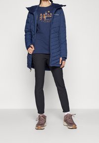Blaue, isolierte Jacke mit Kapuze, kombiniert mit einem marineblauen T-Shirt mit Grafikdruck, schwarzen Hosen und braunen Wanderschuhen. Strukturiertes Material und Reißverschlussdetails.