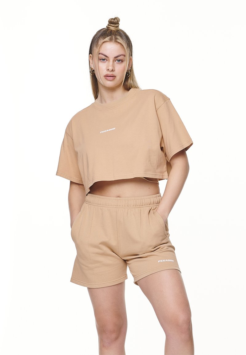Lysbrun cropped T-shirt og matchende shorts lavet af blød bomuld. T-shirten har korte ærmer og et logo, mens shortsene har elastisk talje.