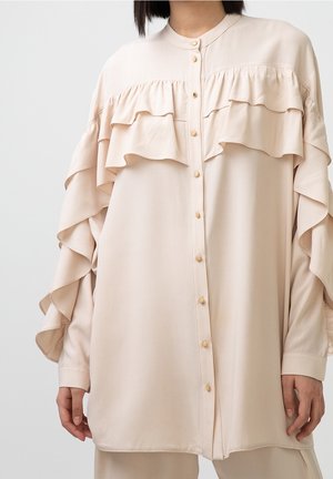 Kobieta w beżowej bluzce zapinanej na guziki, z warstwowymi falbanami na wysokości klatki piersiowej i wzdłuż rękawów, stojąca na tle jednolitego tła.