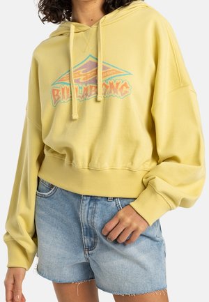 Person trägt einen gelben Billabong Hoodie mit farbenfrohem Logo und hellblaue Jeansshorts mit ausgefransten Rändern.