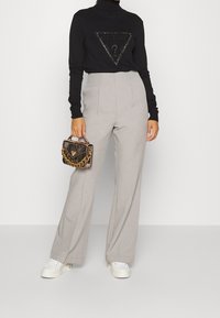 Maglia nera a collo alto con un logo triangolare "GUESS", abbinata a pantaloni grigi chiaro a vita alta e sneakers bianche, che tiene una borsa a motivo fantasia.