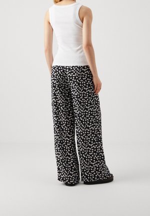 Personne portant un haut blanc sans manches et un pantalon noir à jambes larges avec un petit motif de marguerites blanches, debout devant un fond uni.