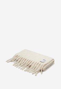 Couverture en laine de couleur blanc crème avec des bords à franges, texture douce, et une petite étiquette d'entretien attachée sur un côté.