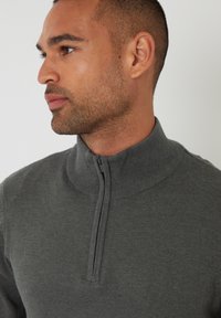 Hombre con cabello corto y barba recortada que lleva un suéter gris con cremallera hasta el pecho, mirando hacia la izquierda sobre un fondo liso claro.