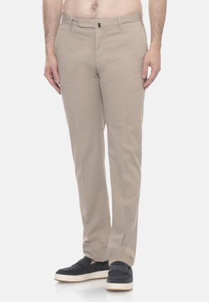 Uomo che indossa pantaloni chino beige modello dritto e sneakers slip-on scure, in piedi davanti a uno sfondo chiaro e uniforme.