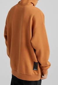 Sudadera naranja de gran tamaño en tejido suave, con cuello alto, hombros caídos y un parche de logo negro en la costura lateral.