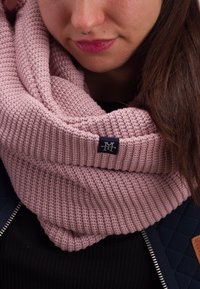 Knitted pink scarf with a textured pattern, featuring a dark blue logo tag. The scarf is wrapped snugly around the neck.