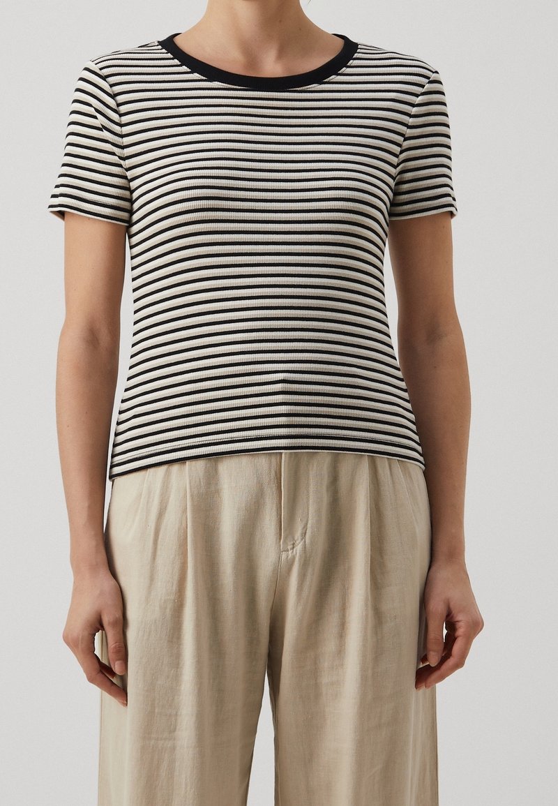 Top court à manches courtes rayé en écru et noir avec une texture côtelée, présentant un col rond et une silhouette ajustée, associé à un pantalon beige.