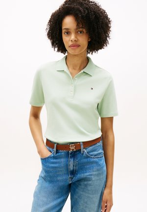 Kobieta o kręconych włosach, ubrana w jasnozieloną koszulkę polo z krótkim rękawem, niebieskie dżinsy i brązowy pasek, z ręką w kieszeni.