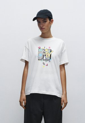 White cotton t-shirt featuring a colorful MTV logo graphic with geometric accents. Crew neckline and short sleeves, paired with dark trousers.