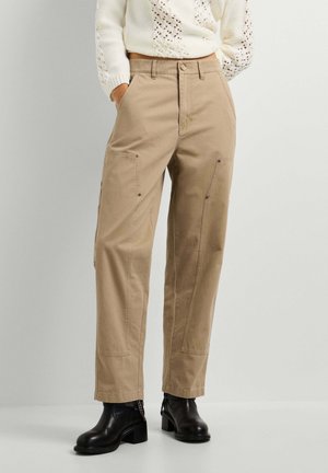 Mujer con pantalones beige de pierna recta con bolsillos, un suéter de punto blanco y botas negras hasta el tobillo, de pie con las manos en los bolsillos.
