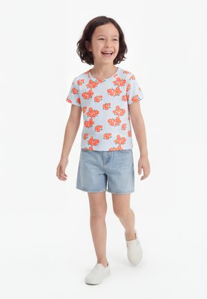 Enfant souriant aux cheveux courts et foncés marchant en avant, portant une chemise florale bleu clair, un short en jean et des chaussures blanches faciles à enfiler sur fond blanc.