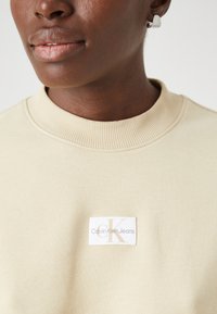 Felpa beige con colletto a coste, con etichetta bianca che riporta "Calvin Klein Jeans" e un logo stilizzato "CK" in oro. Texture morbida.