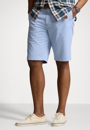 Man met lichtblauwe korte broek, beige sneakers, geruit hemd los gedragen en een polshorloge, met handen in de zakken tegen een effen achtergrond.