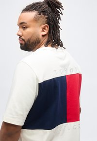 Άνδρας με πλεγμένα μαλλιά που φοράει λευκό κοντομάνικο πουκάμισο με μεγάλο μπλε και κόκκινο τετράγωνο και το κείμενο "Tommy Hilfiger" στην πλάτη.