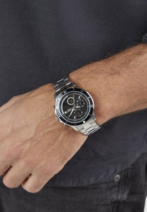 Stainless steel watch with a black dial, luminescent markers, chronograph functions, and a silver metal band, worn on a wrist.