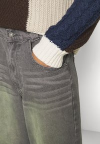 Suéter de punto en una mezcla de colores marrón, marino y crema, con patrones de trenzado. Jeans de mezclilla gris con un acabado de degradado verde deslavado.