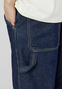 Pantalons en denim bleu foncé avec des coutures jaunes contrastantes, présentant une poche latérale et une texture lisse. La ceinture est partiellement visible.