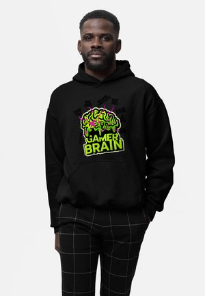 Mężczyzna w czarnej bluzie z kapturem z neonowozielonym i różowym nadrukiem mózgu z napisem "Gamer Brain" oraz w czarnych spodniach w białą kratę.