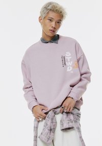 Felpa rosa oversize con grafiche testuali, collo rotondo e polsini a costine, indossata sopra una camicia grigia con colletto. Flanella a quadri annodata in vita.