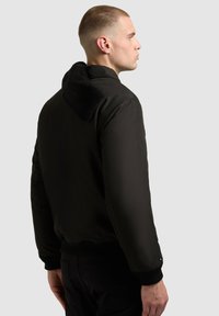 Czarna kurtka bomberka z kapturem podszytym sherpą, ściągaczami na rękawach, gładką fakturą i ukrytym suwakiem. Charakteryzuje się opływowym fasonem.