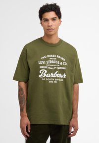 T-shirt en coton vert olive avec des manches courtes, col rond et design graphique en blanc comportant des noms de marque et des dates d'établissement.
