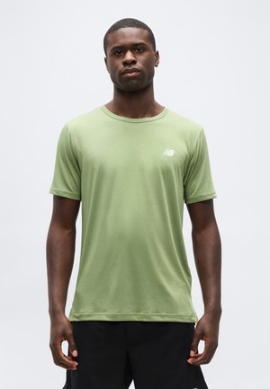 Jeune homme portant un T-shirt New Balance à manches courtes vert clair et un short noir, debout devant un fond gris uni.