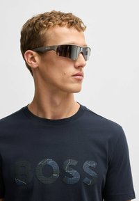 Solglasögon med rektangulär form, mörkt tonade linser och ljusgrå ram. Bärs med en marinblå skjorta med en texturerad "BOSS"-logotyp.